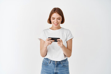 Portrait of young woman watching video on smartphone, holding phone horizontally to play video game, white background