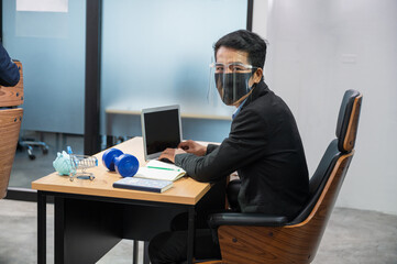 Obraz na płótnie Canvas Asian businessman wearing face shield looking at camera and working with laptop and piggy bank, dumbell, cart and notebook at desk in the office