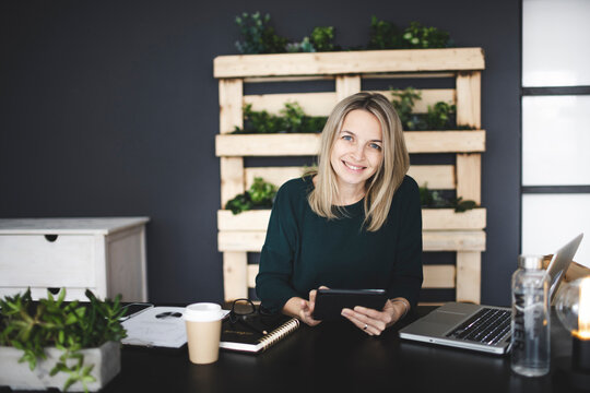 Pretty Young Blond Woman Is Sitting In A Modern, Sustainable Office With Many Green Ecological Plants And Is Working On Her Tablet