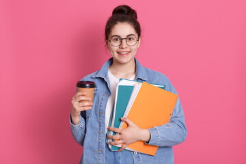 Smiling young European student girl holds folders wears denim jacket. Pleased female with dark hair and hair bun drinks coffee, looks at camera with happy expression, finishes studying.