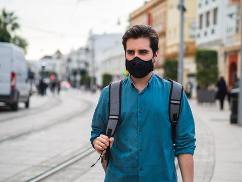 Hombre joven con mochila caminando por la calle vistiendo una mascarilla negra cumpliendo las medidas de seguridad y distanciamiento social de la pandemia del coronavirus