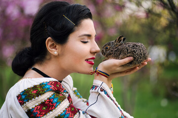 Beautiful young woman posing in romanian traditional costume with a little rabbit in a magnolia...