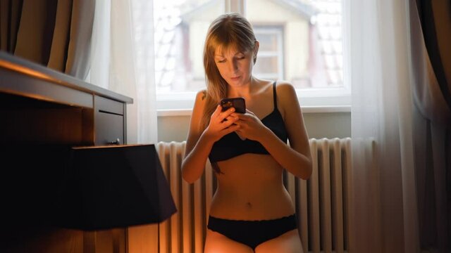 Beautiful young woman in black lingerie texting using mobile phone or cellphone in hotel room