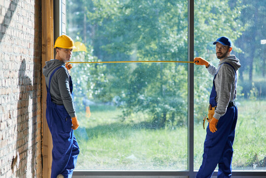 Two positive young male builders wearing blue overalls looking at camera, using measuring tape while working on cottage construction site