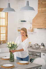 Blonde housewife in jeans putting flowers into the vase