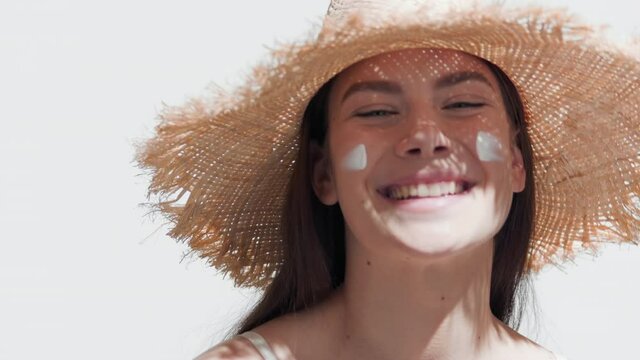 Close-up beauty portrait of cute brown-haired European girl in a straw hat puts spf cream on her cheeks and smiles wide for the camera against white background | Spf cream commercial