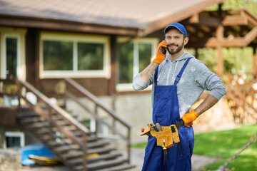 Cheerful young male builder in uniform smiling at camera while talking on the phone, standing outdoors at cottage construction site on a warm sunny day