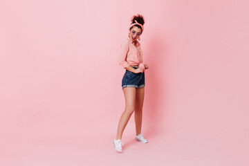 Fototapeta na wymiar Beautiful dark-haired woman in bandage on her head and lilac glasses posing on isolated background. Portrait of girl in white sneakers, shorts and shirt
