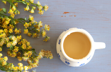 Tea in a mug on wood background with goldenrod herb plant, Solidago virgaurea on a blue background. Treats kidney and urinary tract diseases, improves metabolism.