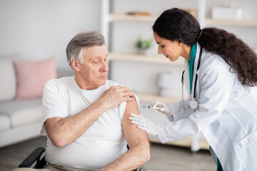 Young doctor making injection of medication or anti-covid vaccine to disabled elderly man in...