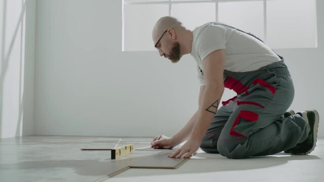 Bald handyman in workwear uses level to check parquet floor
