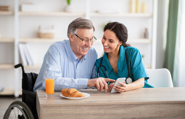Disabled senior man and young nurse using smartphone together, browsing web or watching movie...
