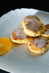 Delicious cottage cheese pancakes with powdered sugar, with an orange on a white plate close-up. Vertical photo