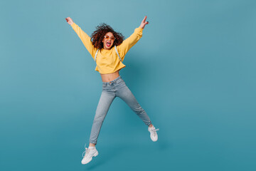 Active girl shows tongue. Woman in yellow sweater and jeans happily jumping on isolated blue background