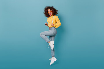 Fototapeta na wymiar Full-length portrait of cheerful curly woman in unusual sunglasses, jumping on blue background. Girl in yellow sweater posing