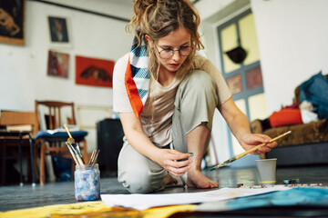 Horizontal image of a pretty female artist sitting on the floor in the art studio and painting on paper with a brush. A woman painter with glasses painting with watercolors in the workshop. - obrazy, fototapety, plakaty