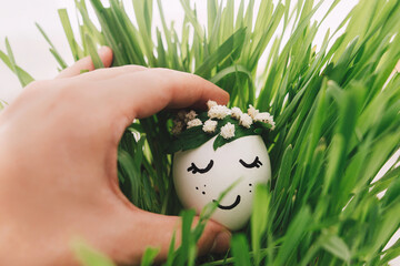Hand holding easter egg with drawn sleeping face in floral wreath in green grass.Easter hunt