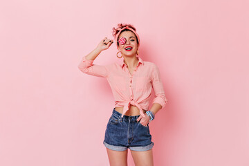 Attractive woman wearing pin-up shirt and headband posing with lollipop on pink background