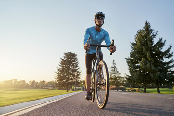 Full length shot of professional male cyclist in sportswear and protective helmet riding road bike in the park at sunset
