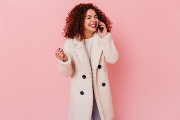 Happy curly brunette girl in white eco-coat happily talking on phone on pink background