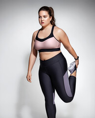 Obraz premium Sporty woman stretching legs before workout. Photo of model with curvy figure in fashionable sportswear on grey background. Sports motivation and healthy lifestyle