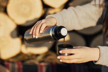 Close up of hands of girl pouring the hot drink from a thermos while sitting near pile of wood logs in forest. Picnic in forest.