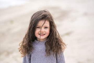 Closeup view portrait of cute pretty white smiling happy little girl with long brown hair standing outdoor on sunny spring sandy beach. Sweet child looking in camera cheerfully