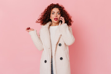 Blue-eyed woman in stylish white coat talking on phone. Photo of curly lady with red lips on pink background