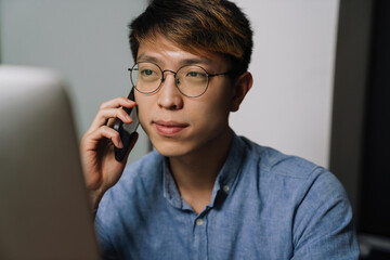 Serious asian guy talking on cellphone while working with laptop