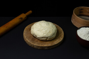 Dough on a dark background. A piece of homemade dough on a wooden board on a black background. The tools for working with the baking dough lie around the dough.