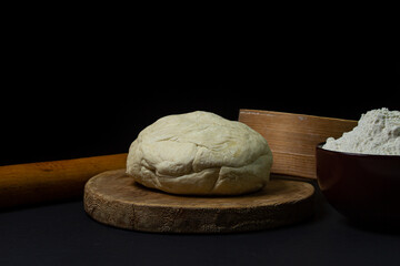 Dough on a dark background. A piece of homemade dough on a wooden board on a black background. The tools for working with the baking dough lie around the dough.