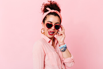 Flirty pin-up girl in glasses licks red lips. Snapshot of woman in massive earrings and pink outfit posing on isolated background