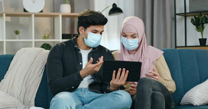 Handsome positive tanned guy in mask and his arabian girlfriend in hijab that is in protective mask too, sitting at home and talking about information they read on tablet device about covid-19