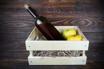Bottle of red wine with cork and two apples in wooden box with paper shavings, still life on brown...