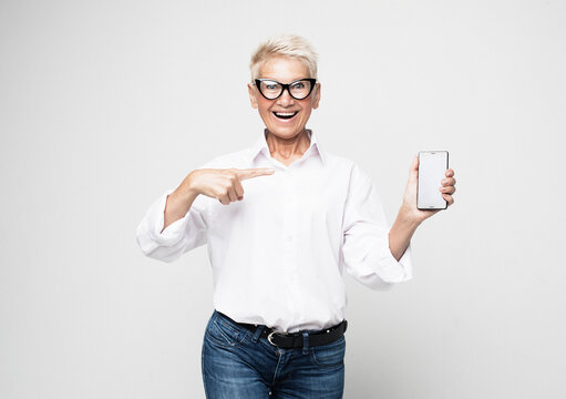 Portrait Of A Senior Elder Smiling Woman Showing Or Presenting Mobile Phone Application And Pointing Finger At The Smartphone Screen On Hand