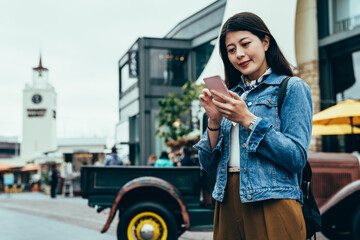 blurred background smiling girl traveling independently is using mobile on the street. young woman standing and looking up information on the internet with phone. authentic lifestyle