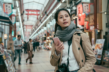 Obraz premium Minami Namba and Shinsaibashi. female tourist standing in local market and searching fresh seafood shop at daytime. young girl carrying camera and holding smartphone look japanese local lifestyle