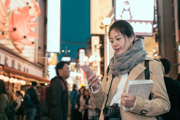 female tourist walking in night shopping street at Dotonbori. young girl photographer holding guide book standing in famous destination for traveler in Osaka japan. woman looking at smartphone screen