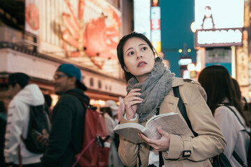 Fototapeta na wymiar Shopping young girl reading guidebook in modern urban. Beautiful woman having fun in city in dotonbori. female tourist standing in crowded searching direction on map in travel book at night city.