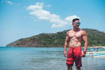 A handsome, fit, tanned, and attractive Filipino man wearing shades and board shorts at the beach....