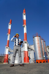 Smiling Manager in Yellow Hardhat Talking on Mobile Phone and Standing Against Power Plant Chimneys and Blue Sky Background