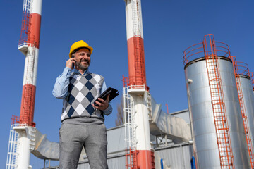 Smiling Businessperson in Yellow Hardhat Talking on Mobile Phone and Standing Against Power Plant Chimneys and Blue Sky Background