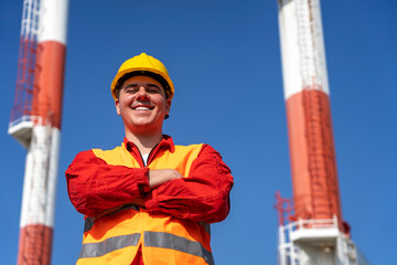Young Worker in Red Coveralls and Yellow Hardhat Standing Against Power Plant Chimneys and Blue Sky Background