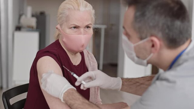 Chest-up over shoulder of aged Caucasian senior blond woman wearing protective face mask, sitting in front of unrecognizable medical worker giving her flu shot on shoulder, lady leaving
