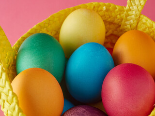 Fototapeta na wymiar Yellow straw basket with multi colored eggs on pink background.