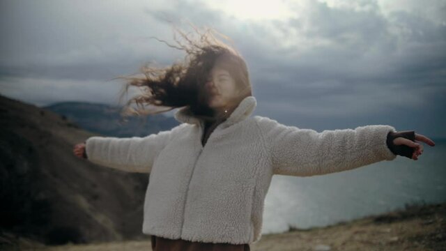Free Happy Young Hiker Woman in warm jaket looking up with raised arms enjoying windy sunny day in the nature breathing fresh air, hair blowing in wind, People Mountains Freedom Concept, Happiness
