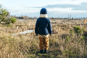 Back view of a child wearing winter clothes standing in front of wild landscape and factories on...