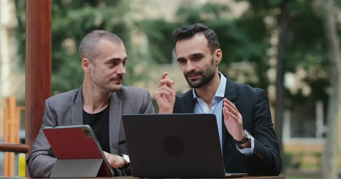 Two positive-minded businessmen discuss the business development prospects in the modern realities conditions, gesticulating while sitting on the summer cafe territory.