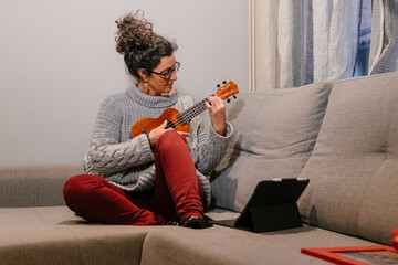 Mujer caucásica de mediana edad sentada en el sofa con ropa casual aprendiendo  a tocar el ukelele por internet a traves de una tablet. - obrazy, fototapety, plakaty