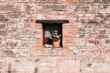 A small window under a wooden lintel decorated with a vase with white and violet flowers in a stone building of a medieval village (Marche, Italy, Europe)